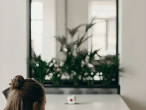 En kvinde med langt, brunt hår arbejder på en bærbar computer ved et hvidt skrivebord i et moderne kontormiljø. Foran hende står en kop kaffe, og i baggrunden ses grønne planter og en stor glasrude, der adskiller kontorområdet.