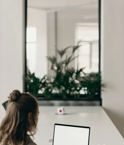 En kvinde med langt, brunt hår arbejder på en bærbar computer ved et hvidt skrivebord i et moderne kontormiljø. Foran hende står en kop kaffe, og i baggrunden ses grønne planter og en stor glasrude, der adskiller kontorområdet.