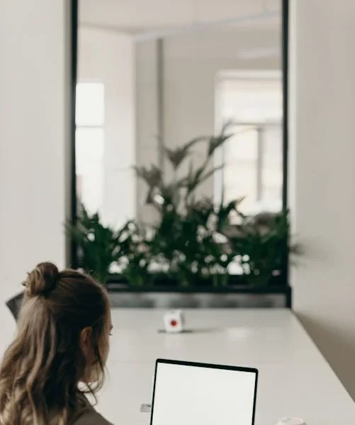 En kvinde med langt, brunt hår arbejder på en bærbar computer ved et hvidt skrivebord i et moderne kontormiljø. Foran hende står en kop kaffe, og i baggrunden ses grønne planter og en stor glasrude, der adskiller kontorområdet.