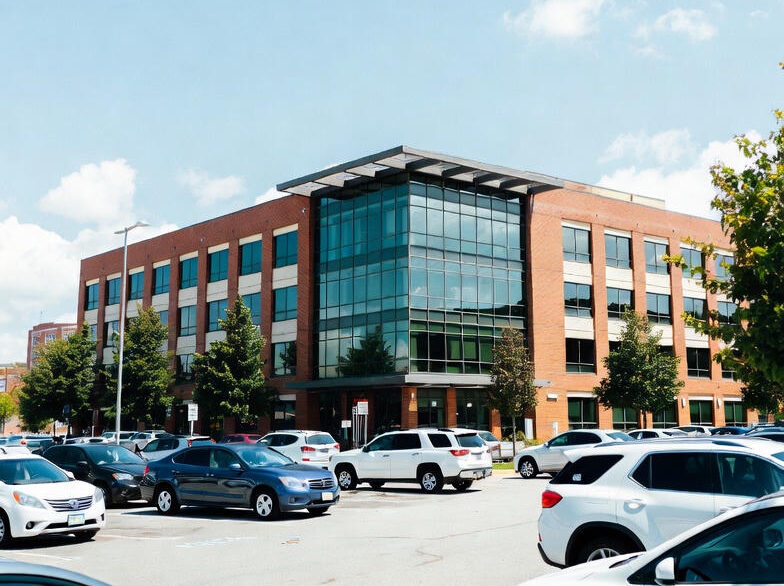 Vid udsigt over en næsten fyldt parkeringsplads på en solrig dag, med mange parkerede biler i forskellige farver, foran et moderne erhvervsbyggeri i røde teglsten med stort centralt glasparti og flere etager. Blå himmel med lette skyer.